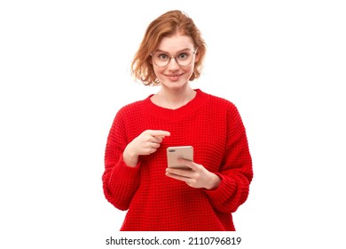 Shocked joyful redhead girl points finger at smartphone blank screen in spectacles and red clothes isolated on white studio background