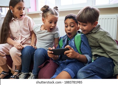 Shocked Kids Using Modern Smartphone With Surprised Emotions And Eyes Wide Open While Sitting At The Break At The Classroom At The School. Children, Science And Education Concept