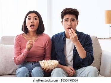 Shocked Korean Couple Watching Horror Movie On TV Eating Popcorn Sitting On Sofa In Living Room At Home. Chinese Family Watches Television, Scary Films For Weekend Concept