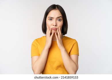 Shocked korean girl touching her face, looking concerned at camera, standing in yellow t-shirt over white background