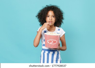 Shocked Little African American Kid Girl 12-13 Years Old In Striped Clothes Isolated On Blue Wall Background Studio. People In Cinema, Lifestyle Concept. Watching Movie Film, Hold Bucket Of Popcorn