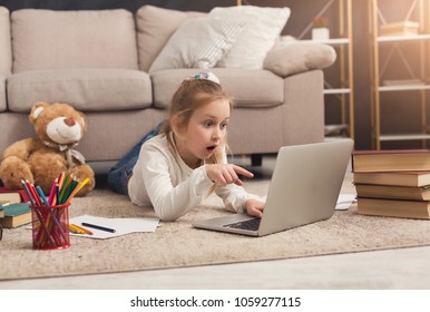 Shocked little casual girl watching movie. Surprised female kid lying on the floor, home alone, watching forbidden scary movies, pointing on laptop - Powered by Shutterstock