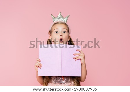 shocked little girl in crown holding book while looking at camera isolated on pink