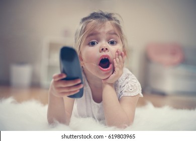  Shocked Little Girl Watching TV Lying On Floor With Remote Control In Hand.