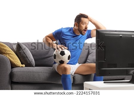 Shocked male athlete sitting on a sofa, holding a football and watching a match on telly isolated on white background