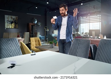 Shocked male customer looking at pricetag, cannot believe high steep price in store. Front view of perplexed man inquiring about cost of furniture, with interior goods on background. Emotions concept.
