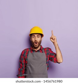 Shocked male industrial worker wears yellow hardhat, apron, points upwards, shows blank space above, isolated on purple background. Mechanical worker or repairman in uniform busy with hard work. - Powered by Shutterstock