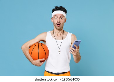 Shocked Man Basketball Player With Thin Skinny Body Sportsman In Headband Shirt Hold Ball Using Mobile Cell Phone Typing Sms Message Isolated On Blue Background. Workout Gym Sport Motivation Concept