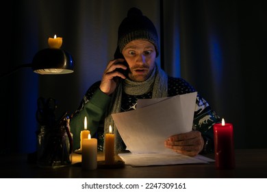 shocked man in candle light looking at high bills for electricity and gas heating in cold dark room at home. energy crisis, household expenses and inflation concept