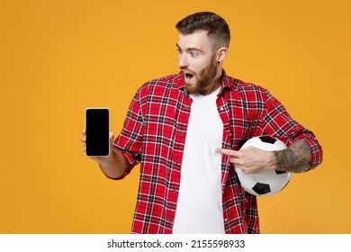 Shocked Man Football Fan In Red Shirt Cheer Up Support Favorite Team With Soccer Ball Point Finger On Mobile Phone With Blank Empty Screen Isolated On Yellow Background. People Sport Leisure Concept