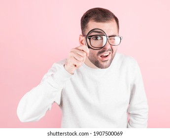 shocked man with glasses with a funny expression in a sweatshirt looks through a magnifying glass on a pink background