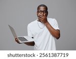 shocked man holding laptop, surprised expression, casual white t shirt, modern glasses, gray background, technology concept, emotional response to online news, digital communication