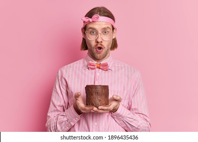 Shocked Man Nerd With Bob Hairstyle Wears Big Transparent Glasses Holds Small Cake With Burning Candle Celebrates Successful Finishing Of University Wears Shirt And Bowtie Isolated Over Rosy Wall