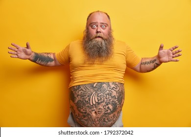 Shocked Man With Thick Beard Raises Arms, Tells About Very Huge Size Of Something, Impressed By What He Sees, Has Big Tattooed Stomach, Wears Yellow T Shirt. Chubby Fat Male Model Gestures Emotionally