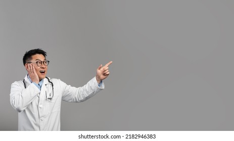 Shocked Mature Chinese Man Doctor In White Coat, Glasses With Stethoscope Shows Finger On Empty Space, Isolated On Gray Background, Studio. Health Care, Huge Medical Ad And Offer And People Emotions