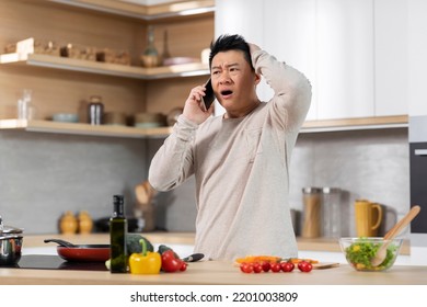 Shocked Middle Aged Asian Man In Casual Outfit Standing Next To Kitchen Table At Home, Cooking Healthy Meal And Talking On Mobile Phone, Touching His Head And Looking At Copy Space