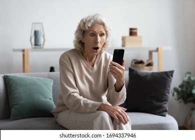 Shocked Middle Aged Grey Haired Woman Resting On Sofa, Looking At Smartphone Screen, Reading Message, Surprised By Unbelievable News. Excited Mature Granny Received Online Lottery Win Notification.