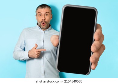 Shocked Middle Aged Male Showing Mobile Phone With Huge Blank Screen Pointing Finger Posing Looking At Camera Over Blue Studio Background. Check This Application, Great Offer Concept. Mockup