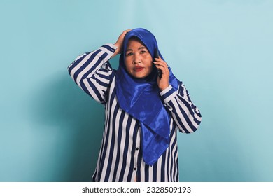 A shocked middle-aged Asian woman in a blue hijab and a striped shirt is talking on the phone with a surprised expression, hearing big news. She is isolated on a blue background - Powered by Shutterstock