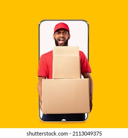 Shocked Millennial Black Guy Delivery Man Gives Boxes From Blank Screen Of Huge Smartphone, Isolated On Orange Background, Studio. Application For Ordering And Delivery Of Purchases. Good Offer And Ad