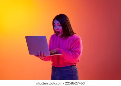 Shocked news  Young girl in pink sweater emotionally looking laptop over gradient orange background in neon light  Freelancer  Concept emotions  facial expression  youth  inspiration  sales  ad