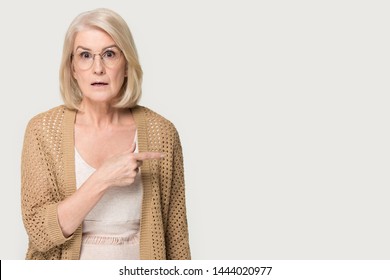 Shocked Old Lady Showing To Left At Copy Space Blank Place. Confused Elderly Woman Attracts Attention, Advice, Help, Invite Concept. Headshot Of Senior Female In Studio Isolated On Gray Background