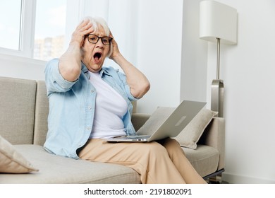 A Shocked Old Lady Is Sitting On A Beige Sofa And Screaming Loudly With Her Mouth Wide Open, Clutching Her Head With Her Hands And Holding A Laptop On Her Lap