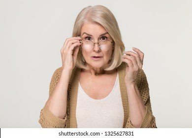 Shocked Old Mature Woman In Disbelief Lowering Glasses Looking At Camera, Surprised Stunned Amazed Senior Mid Aged Lady Grandma Peering Feeling Astonished Isolated On Grey White Studio Background