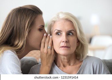 Shocked Older Woman Listening To Young Female Whispering In Ear, Friend Gossiper Telling Secret Rumors Or Unbelievable News To Surprised Senior Lady, Old Mother And Adult Daughter Gossiping Concept