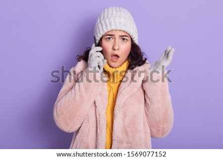 Shocked outraged young woman standing isolated over lilac background in studio, gossiping over mobile phone, putting her smartphone close to ear, opening her mouth widely. Breaking news concept.