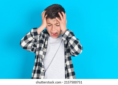 Shocked panic caucasian kid boy wearing plaid shirt over blue background holding hands on head and screaming in despair and frustration.