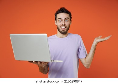 Shocked Perplexed Young Bearded Man Wearing Casual Basic Violet T-shirt Standing Working On Laptop Pc Computer Spreading Hands Isolated On Orange Color Background Studio Portrait. Tattoo Translate Fun