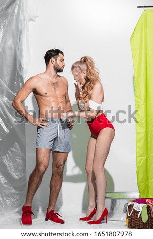 shocked pin up woman touching swim trunks of man and covering mouth on white