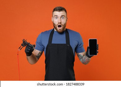 Shocked professional tattooer master artist man in t-shirt apron hold machine black ink jar equipment for making tattoo art on body mobile phone with blank empty screen isolated on brown background