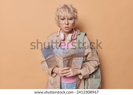 Shocked puzzled female tourist navigates world map in her hands exudes spirit of summer explorer dressed in casual clothes carries backpack isolated over brown background. Quest for adventure