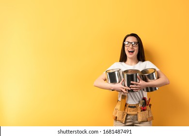 Shocked Repairwoman Holding Paint Cans And Looking Away On Yellow Background