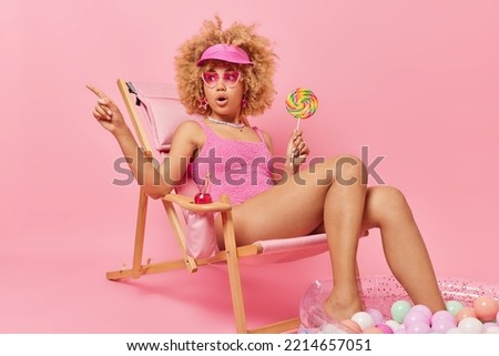 Shocked scared curly haired woman wears heart shaped sunglasses and swimsuit holds big sweet candy on stick points into distance poses on deck chair keeps legs in inflated pool isolated on pink wall