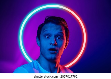 Shocked, scared. Young man's portrait isolated on blue studio background in multicolored neon light with geometric luminescent shape cirlce. Concept of human emotions, facial expression, ad, fashion.