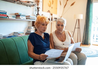 Shocked senior lesbian couple holding bills looking at laptop on couch - Powered by Shutterstock