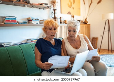 Shocked senior lesbian couple holding bills looking at laptop on couch - Powered by Shutterstock