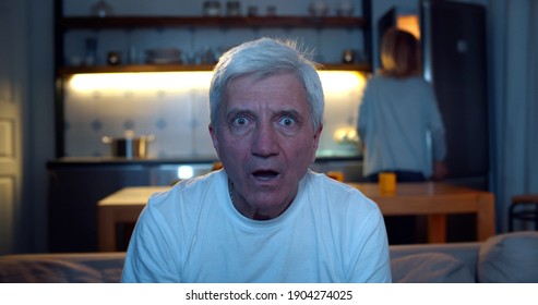 Shocked Senior Man Sitting On Sofa Watching Interesting Tv Show. Close Up Portrait Of Astonished Aged Male With Open Mouth Watching Tv News Sitting On Couch With Wife Cooking On Background