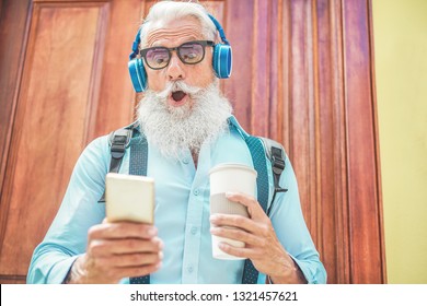 Shocked senior man using smartphone while going out from home listening music and drinking coffee at morning time - Technology, trendy lifestyle,  and joyful elderly lifestyle concept - Focus on face - Powered by Shutterstock