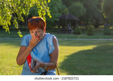 Shocked Senior Red Haired Bankrupt Woman With Empty Wallet. Bankruptcy. Old Woman Hands Holding Open An Empty Wallet. Finance Problems. Embarrassed Woman Looking For Money In Her Wallet In Summer Park