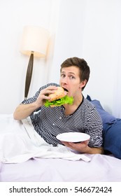 Shocked Single Man Eating Hamburger While Lying In Bed. Handsome Man Looking At Camera And Having Breakfast In Morning At Home.