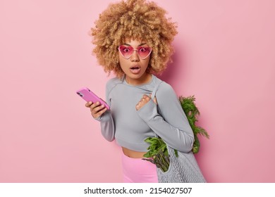 Schockierte schlanke Frau kehrt zurück, nachdem Einkaufstasche voll mit frischem grünem Gemüse trägt, verwendet Handy-Bestellungen Lebensmittelgeschäft online genießt Marktpreis in Aktionsbekleidung gekleidet auf rosafarbenem Hintergrund