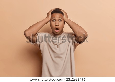 Shocked speechless adult man keeps hands on head mmouth opened stares with omg expression at camera realizes something horrible dressed in casual t shirt isolated over brown background. Omg concept