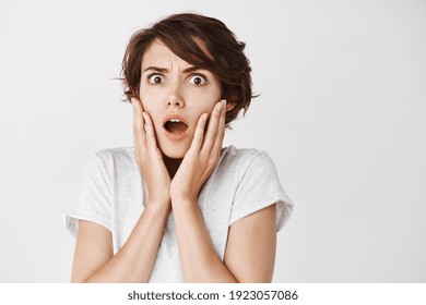 Shocked and startled caucasian woman gasping and frowning, looking at something terrible, standing on white background in casual t-shirt.