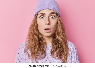 Shocked startled Caucasian woman has eyes popped out looks with amazement holds breath wears purple hat and knitted jumper isolated over pink background. Human reactions and emotions concept