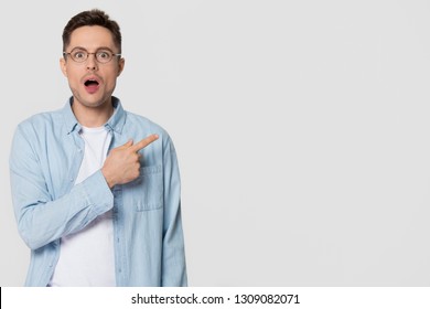 Shocked stunned young man nerd pointing finger aside at blank copy space advertising unbelievable presentation, astonished surprised guy looking at camera isolated on white grey studio background