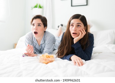 Shocked And Suprised Young Women In Pajamas Resting In The Bedroom While Watching An Action Mystery Movie With A Bowl Of Chips